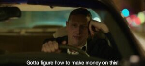 Man driving in a car. subtitles say "gotta figure how to make money on this!"