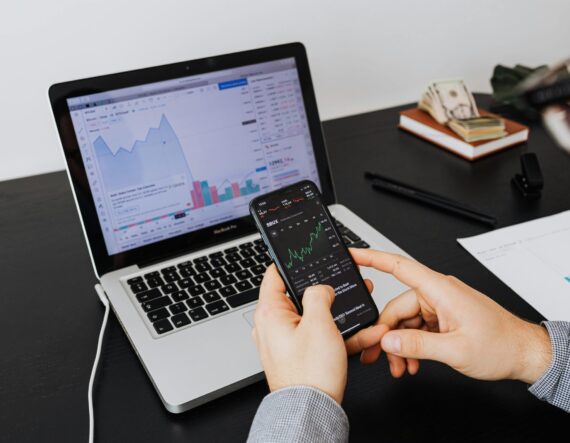 hands holding smartphone looking at the stock market in from of a laptop with more stock graphs on the screen